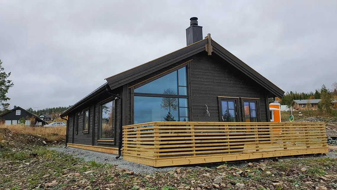Mørk trekledd hytte med store vindusflater og terrasse med gjerde, omgitt av natur og andre hytter.