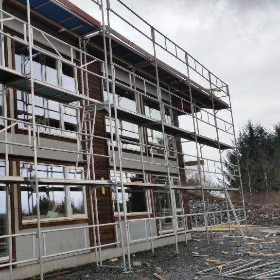 Bygning under oppføring med stillas foran fasade med store vinduer og mørk trekledning.