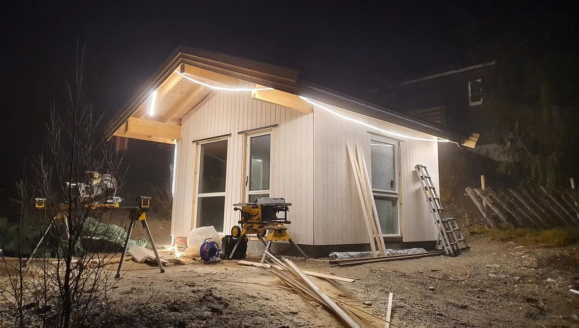 Lite bygg under oppføring med arbeidslys montert i takutstikk og byggeutstyr plassert rundt.