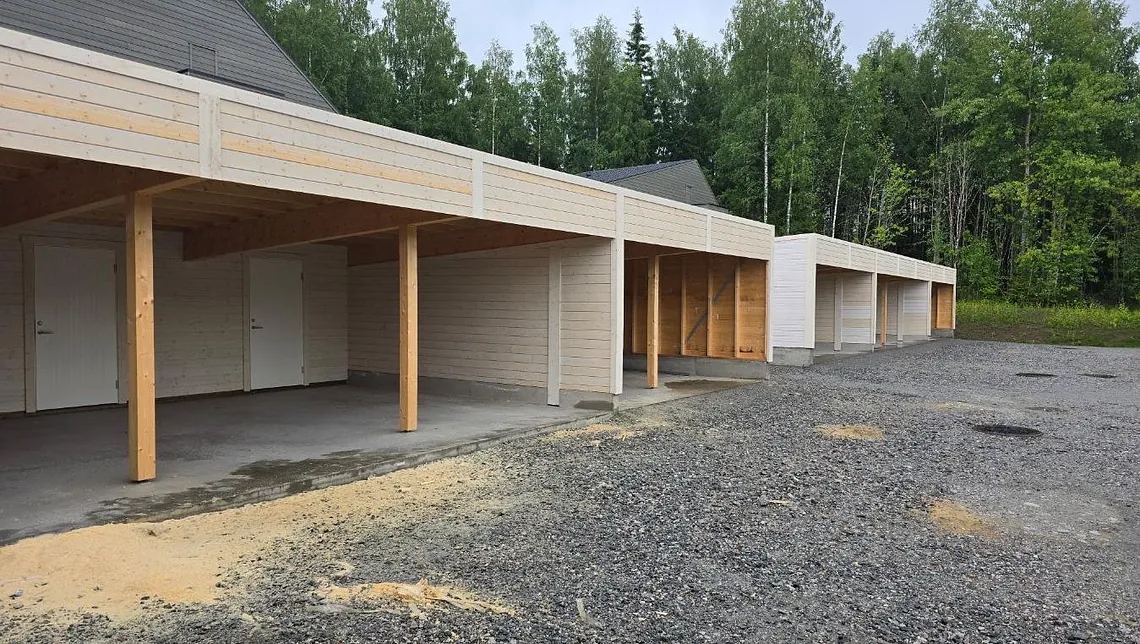 Åpen carport i lyst treverk med boder og støpt dekke, omgitt av grus og skogkledd bakgrunn.