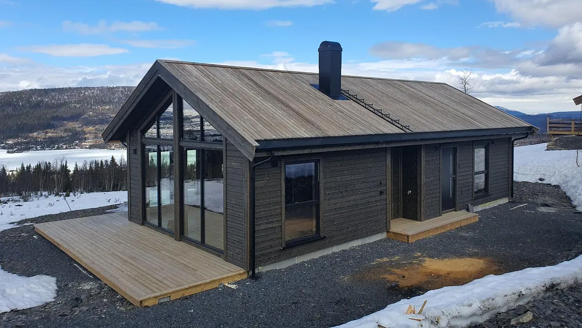 Hytte i mørkt treverk med store vindusflater og terrasse, omgitt av snø og utsikt over fjell og innsjø.