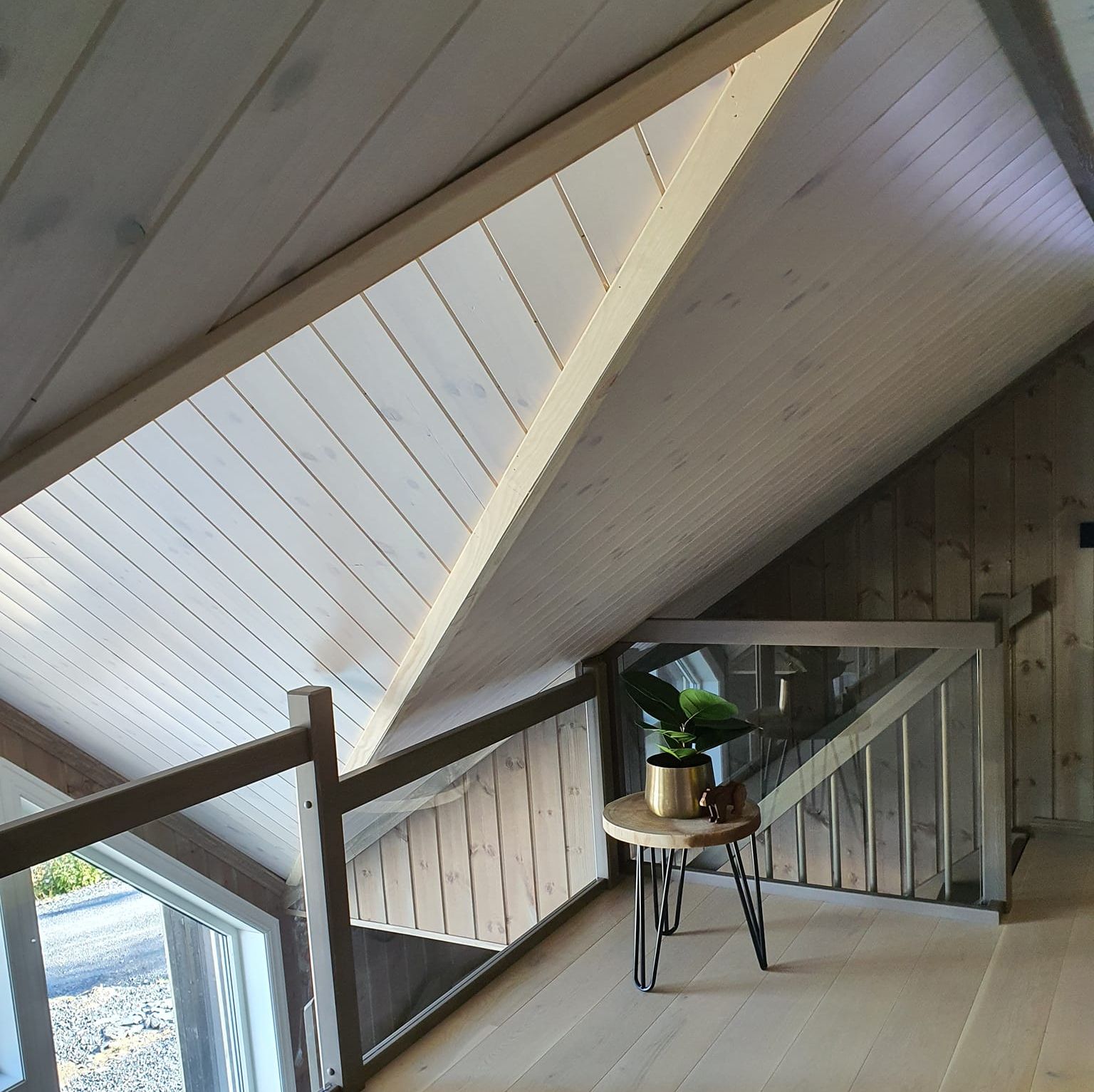 Lys og moderne loftsstue med skråtak, store vinduer, glassrekkverk og lite bord med plante.