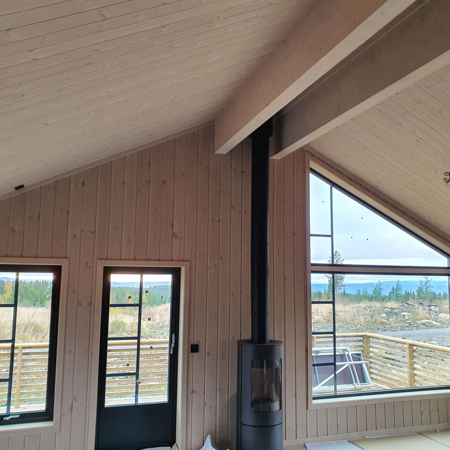 Stue med skråtak, store vinduer og svart vedovn, med utsikt til skog og fjell.