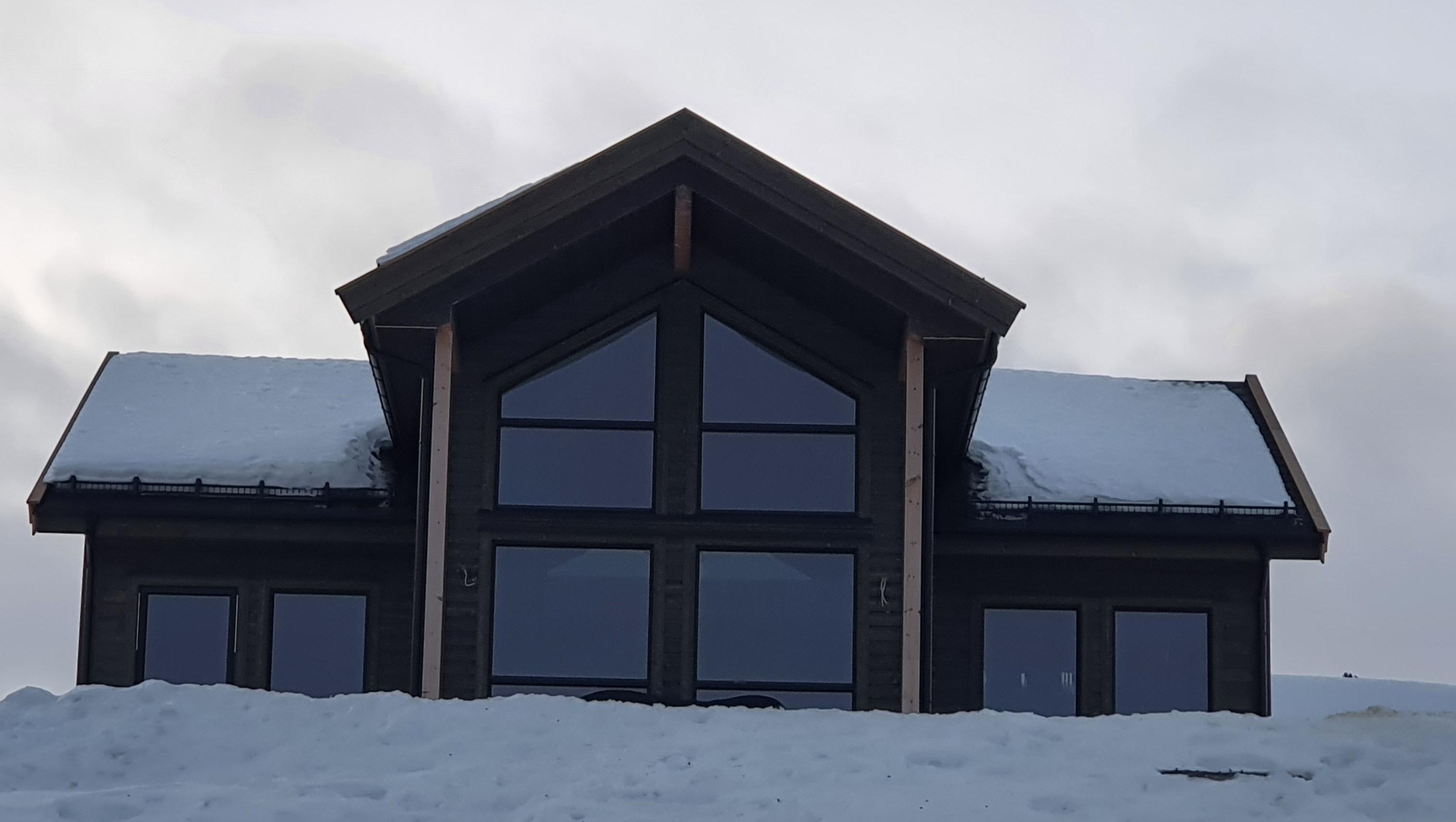 Fasadebilde av hytte med store trekantede vinduer, mørk kledning og snø på tak og terreng.
