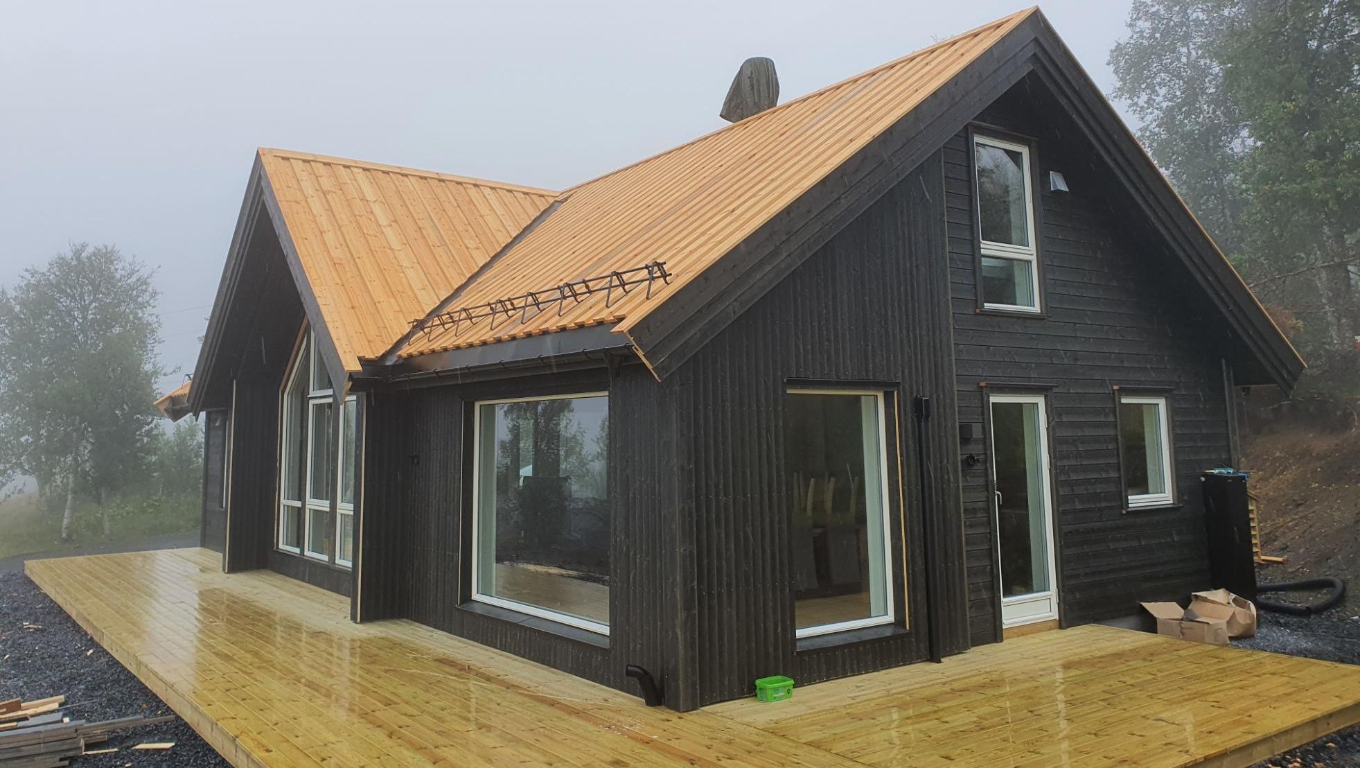 Moderne hytte med mørk fasade og nylagt tretak, omgitt av tåke og våt treterrasse.
